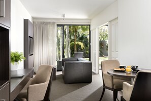 A modern hotel room with a sofa, chairs, a dining table with breakfast items, a potted plant, and a view of palm trees through the glass door.