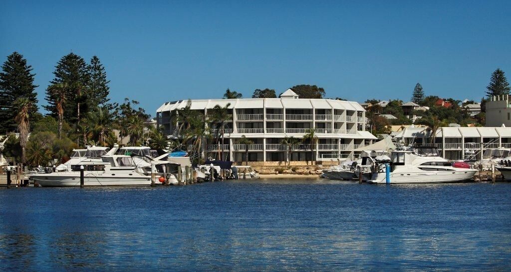 A marina with multiple boats docked, a large white building with balconies, and surrounding greenery.