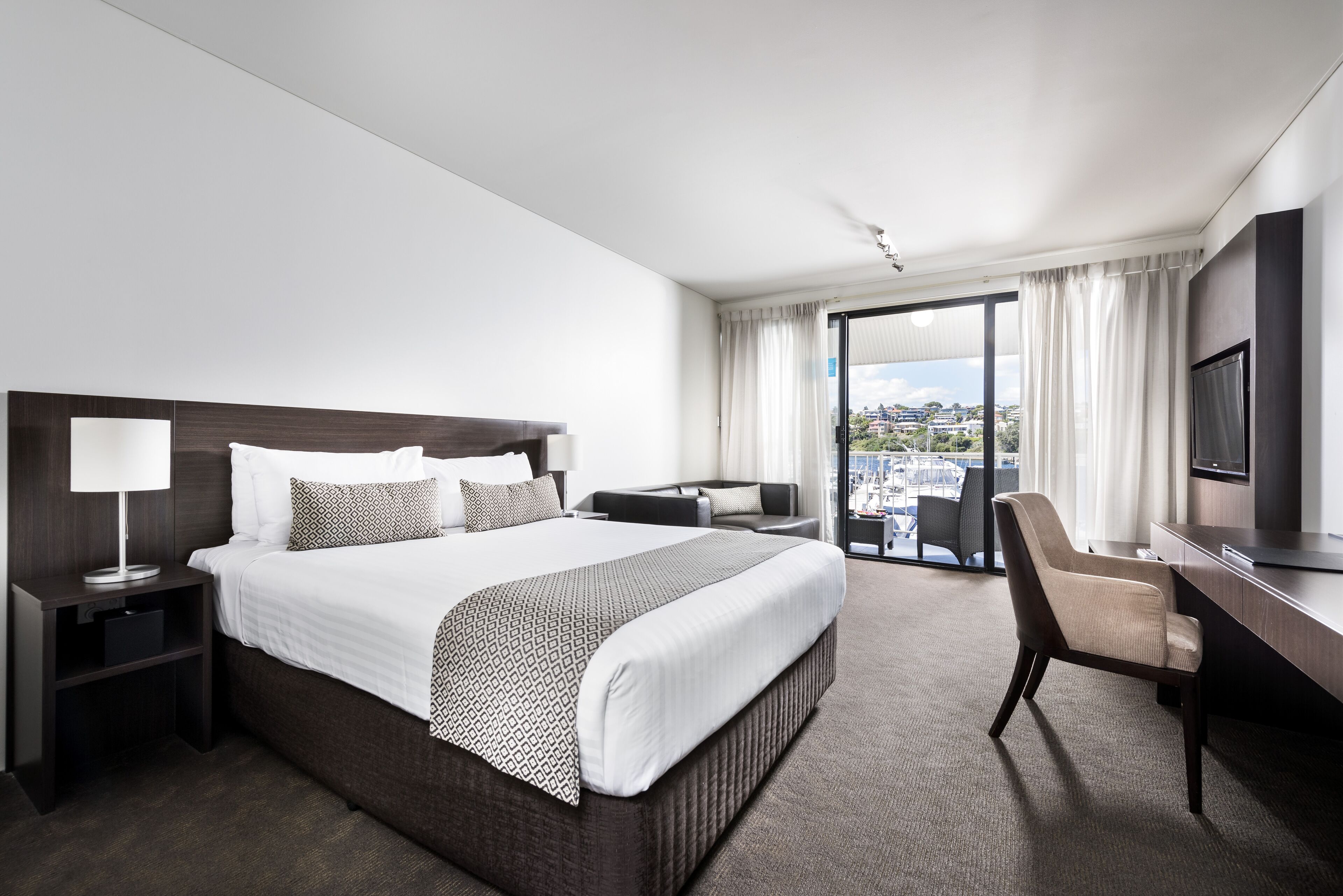 A modern hotel room with a large bed, a desk, a chair, a TV, and a balcony with a city view.