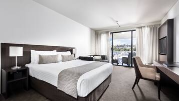 A modern hotel room with a large bed, a desk, a chair, a TV, and a balcony with a city view.