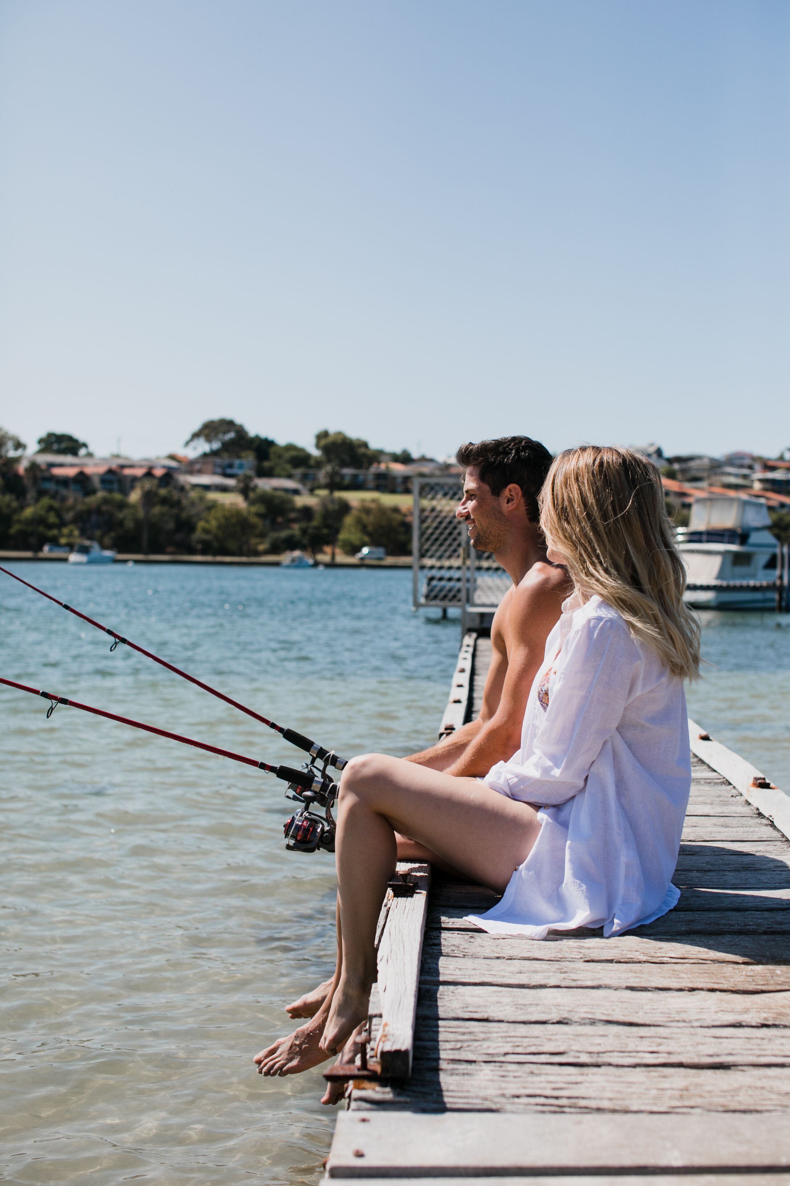 Two people sitting on a wooden dock by a body of water, with fishing rods in hand.