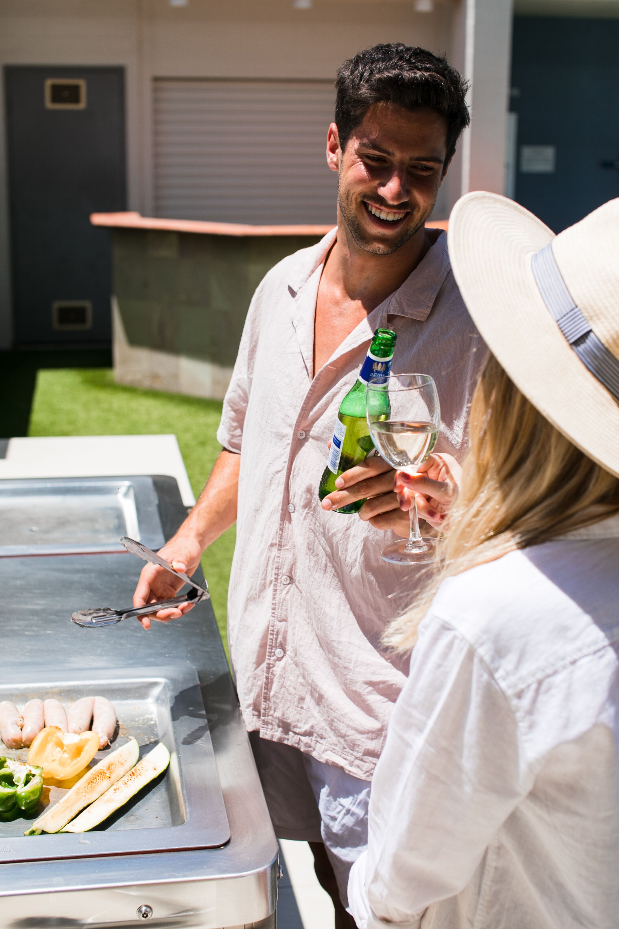 A man holding a bottle and a glass, standing next to a grill with sausages and vegetables.
