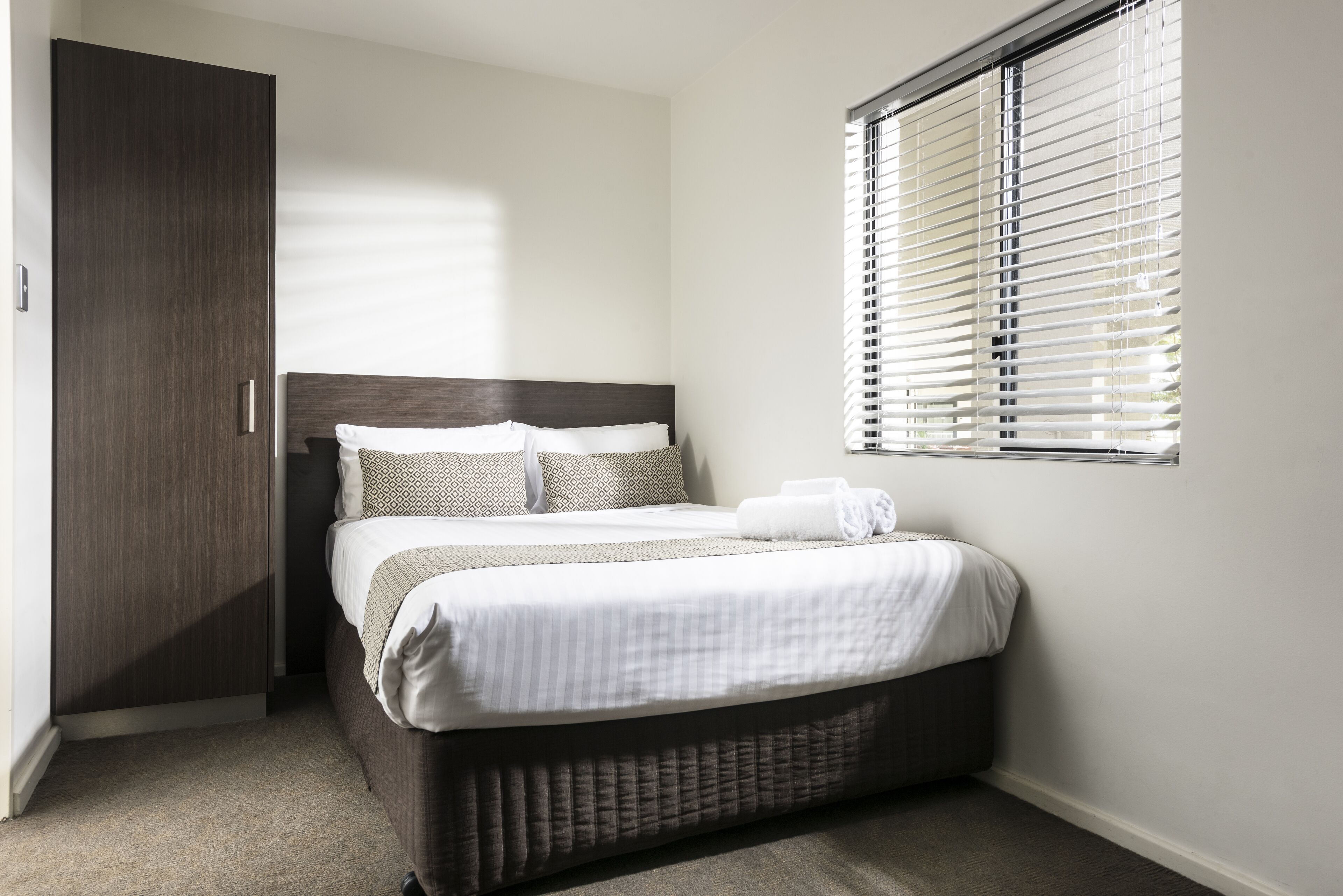 A neatly made bed with a dark headboard, a window with blinds, and a wardrobe.