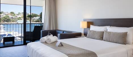 A hotel room with a large bed, a desk, a chair, a balcony with a view of the sea, and a lamp.