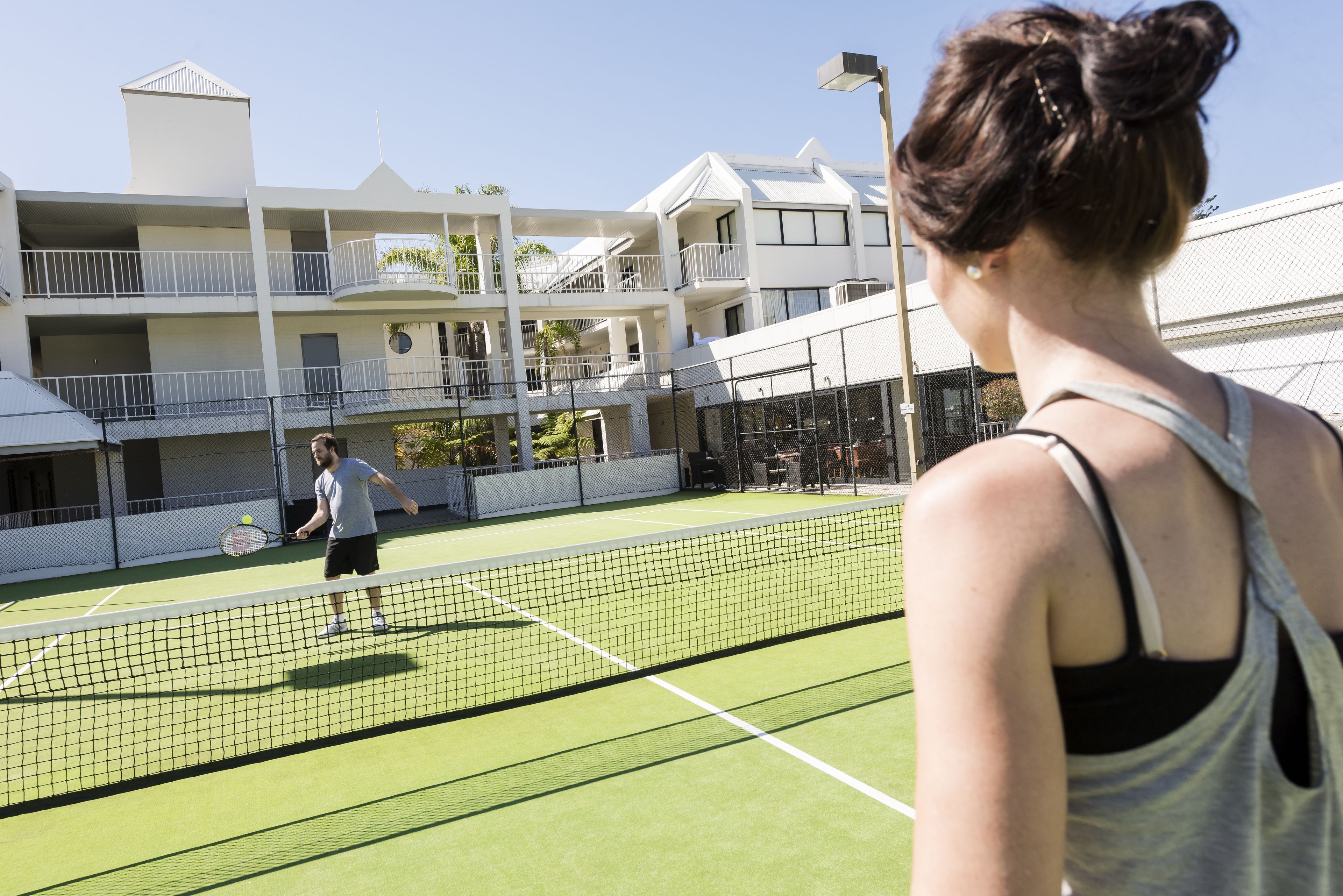 Un court de tennis avec un filet au milieu, entouré d’un bâtiment doté de balcons et d’une clôture.