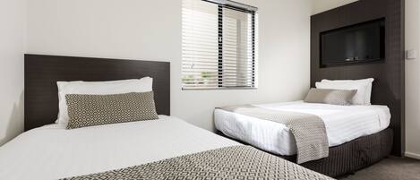 Une chambre d’hôtel avec deux lits, une grande fenêtre avec stores, un téléviseur à écran plat fixé au mur et un sol recouvert de moquette.