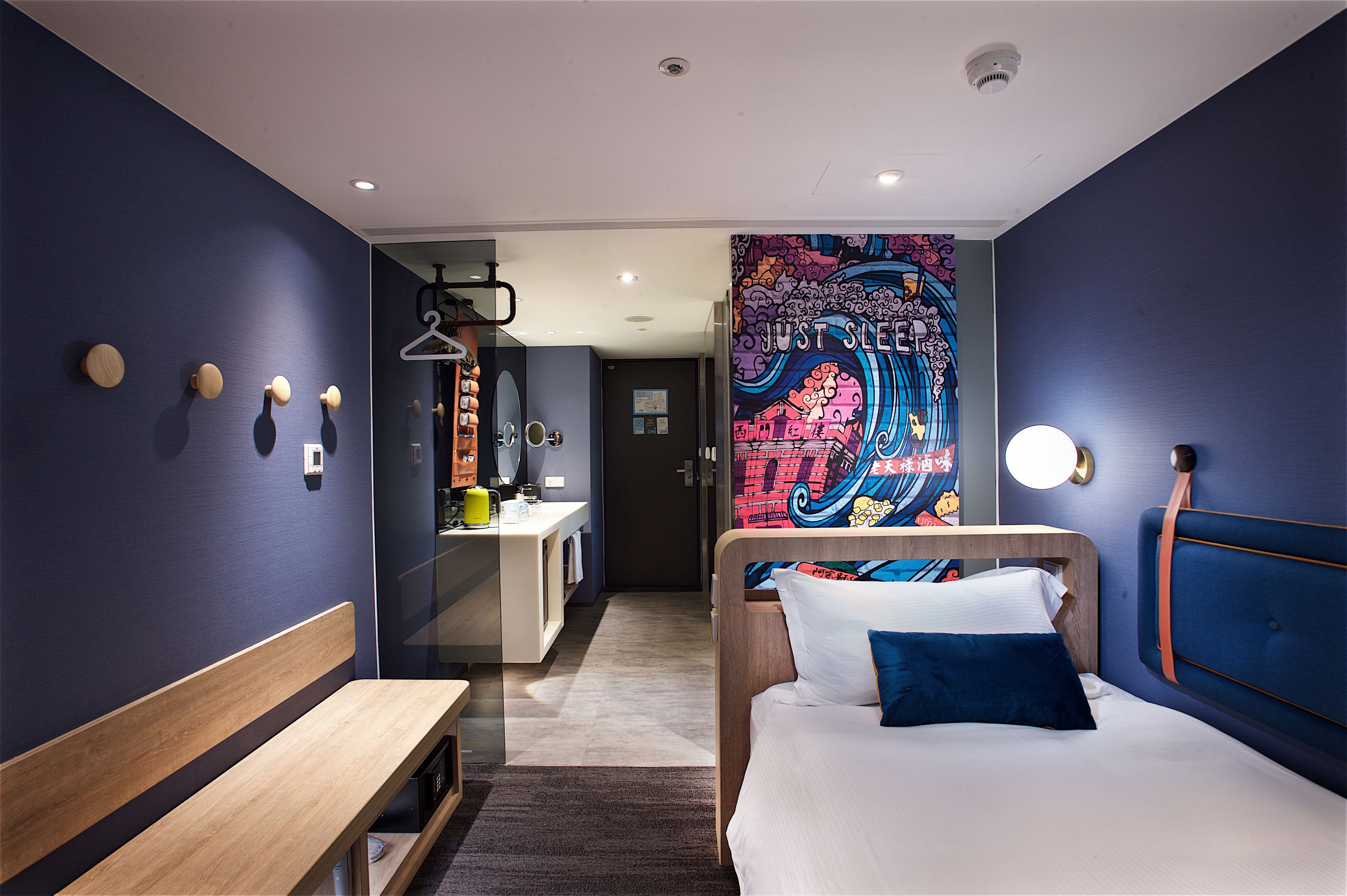 A modern hotel room with a large bed, a wooden bench, a small desk, and a colorful mural on the wall.