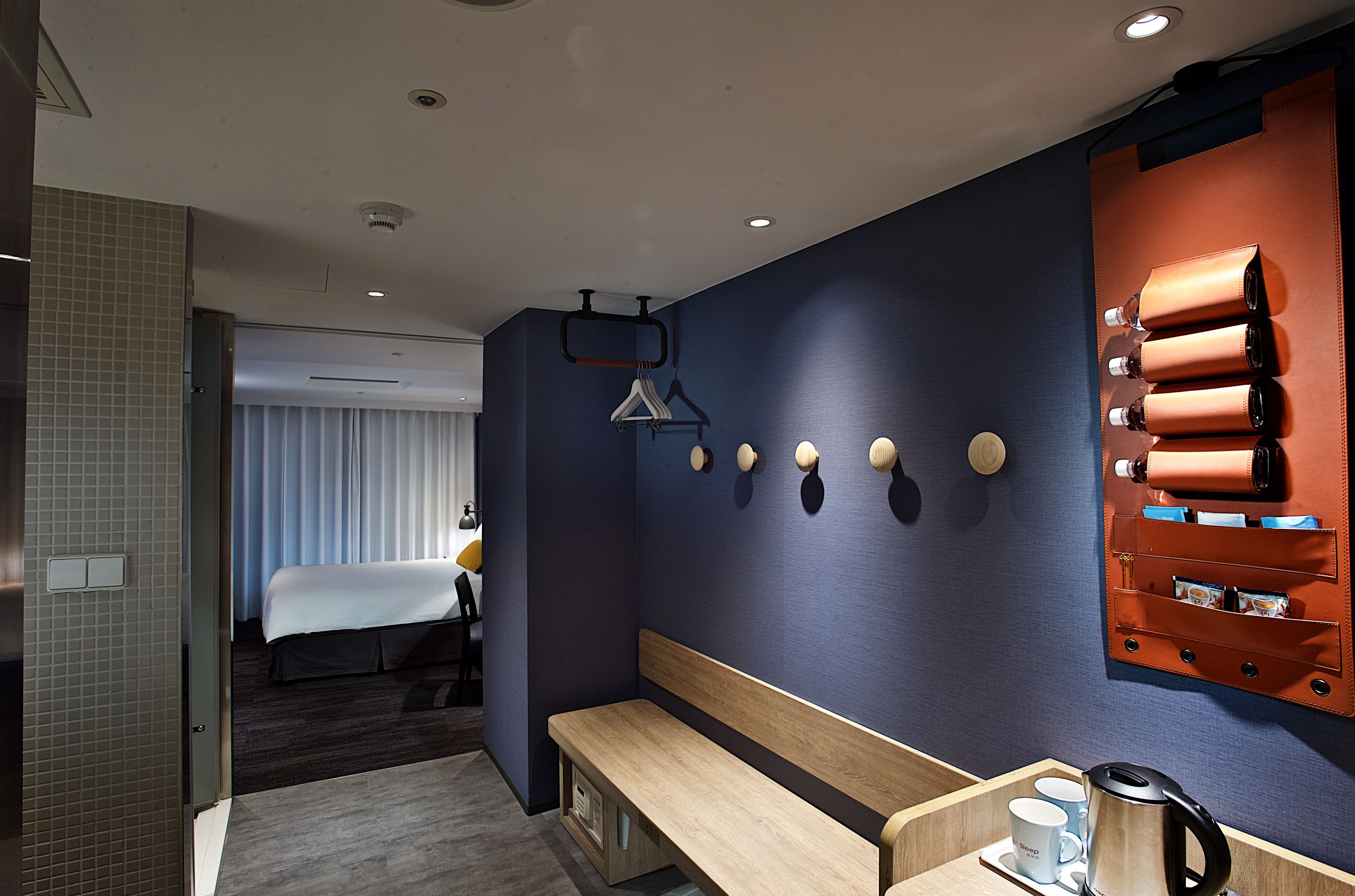 A hotel room with a bed, a bench, a wall-mounted coat rack, and a leather storage unit.