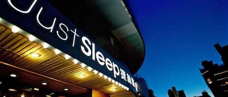A hotel sign with illuminated 'Just Sleep' branding, a curved roof, and a cityscape in the background.