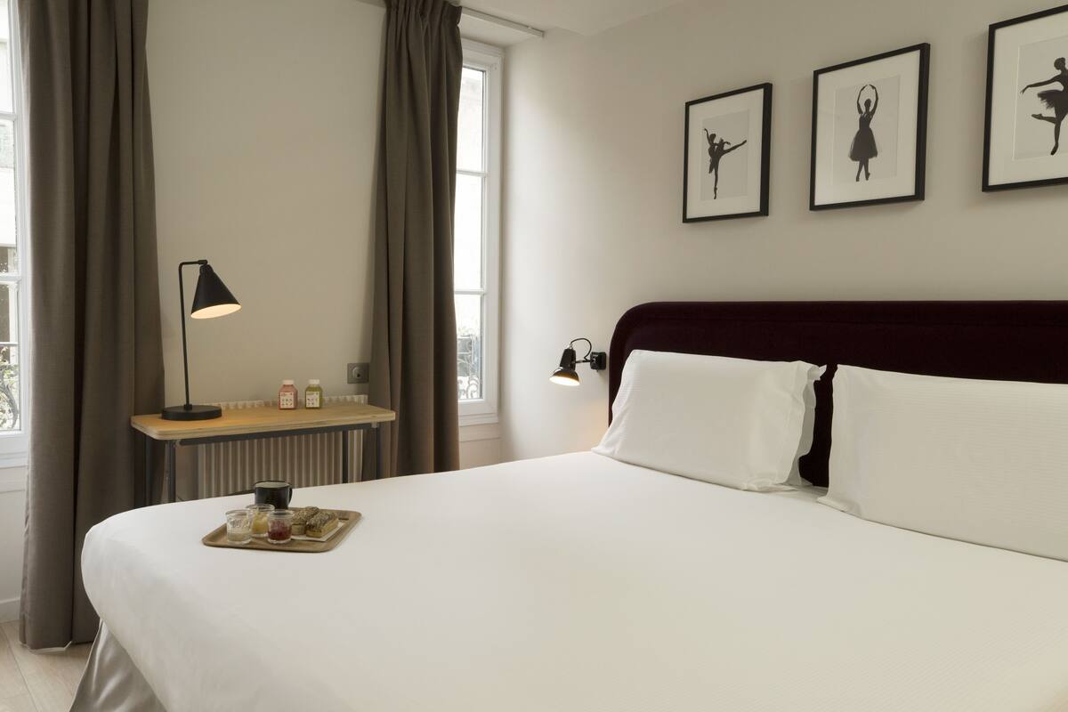 A hotel room with a large bed, a bedside table with a lamp, a tray of snacks, and framed pictures on the wall.