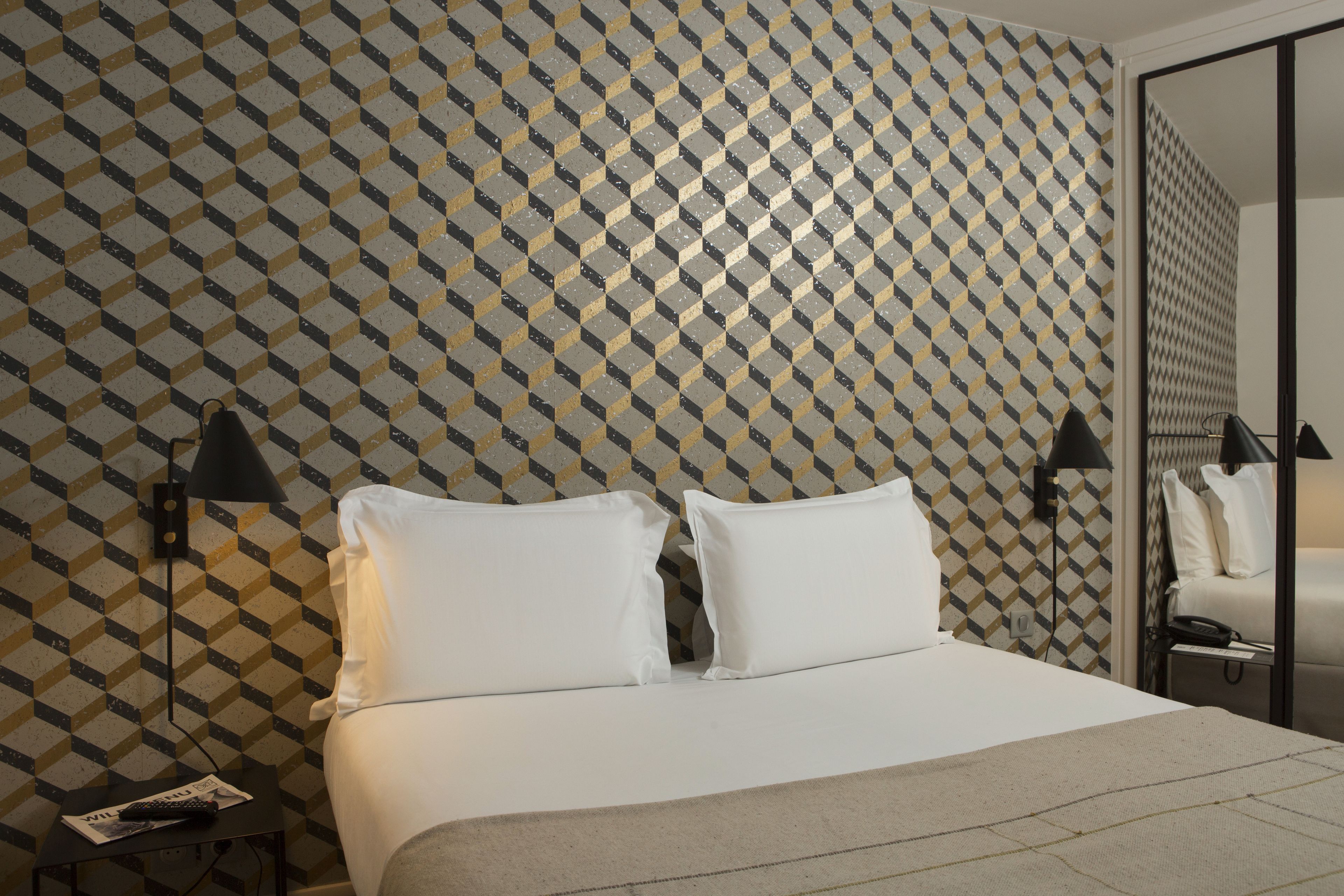 Une chambre d’hôtel avec un papier peint à motifs géométriques, un lit avec du linge de maison blanc, deux lampes de chevet et une table de chevet avec une télécommande.