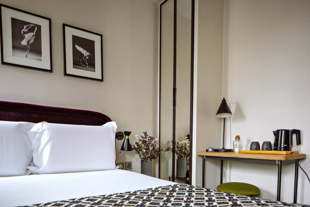 A hotel room with a bed, a desk with a lamp, a chair, and two framed pictures on the wall.