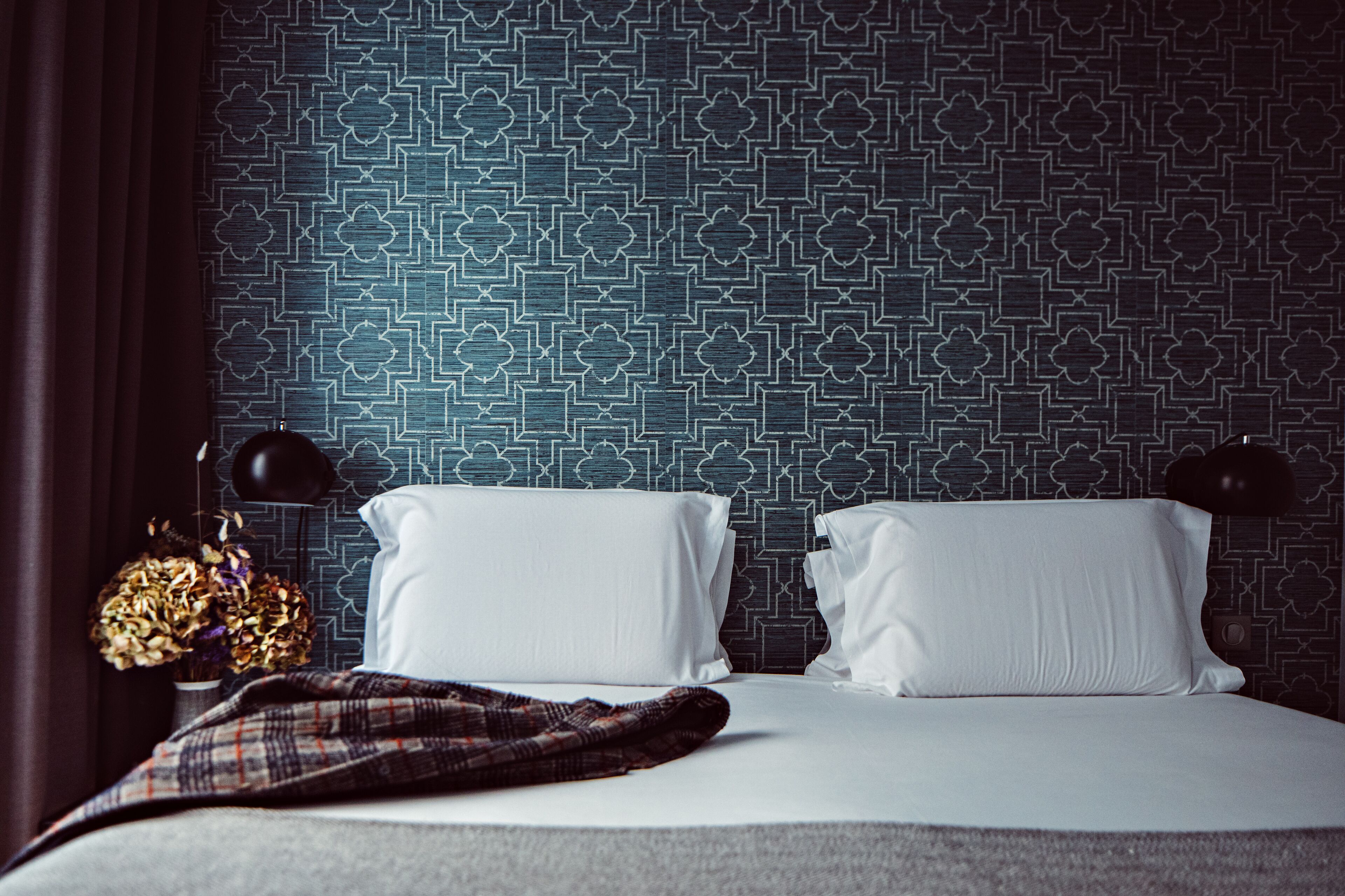 A hotel room with a patterned wall, two white pillows, a plaid blanket, and a vase with dried flowers.
