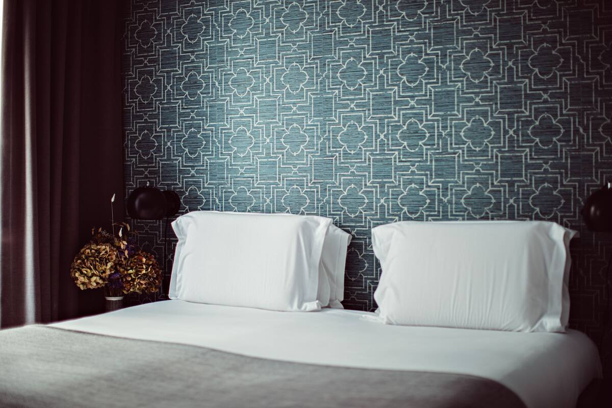 A hotel room with a patterned blue wall, a bed with white linens, and a vase with dried flowers on the nightstand.