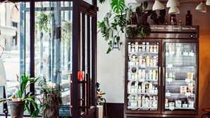 A restaurant interior with a wooden table, chairs, a patterned floor, a refrigerator stocked with beverages, and potted plants. - Monsieur Helder (Paris)