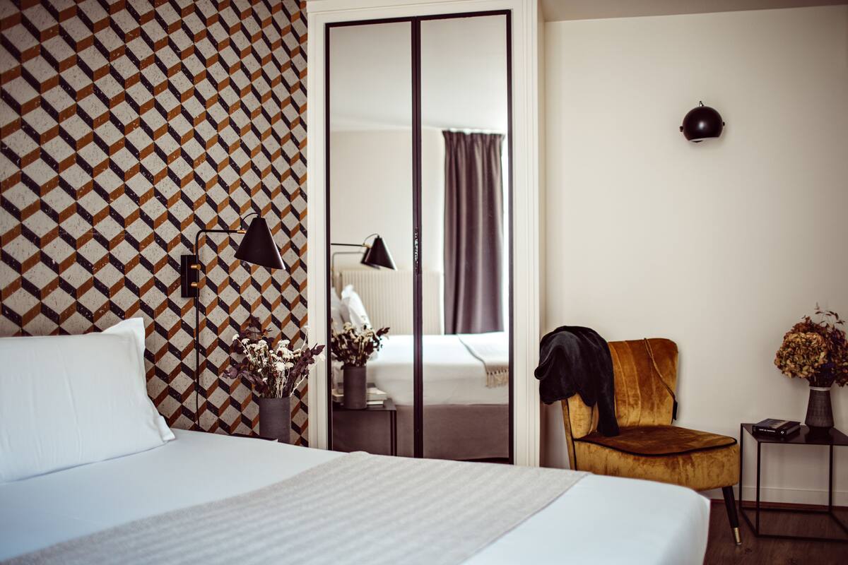 A hotel room with a bed, a chair, a nightstand, a lamp, a mirror, and a patterned wall.