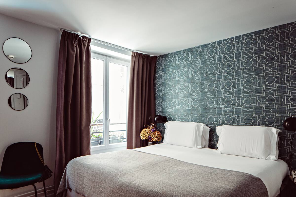 A hotel room with a large bed, a chair, a window with curtains, and a patterned wall.