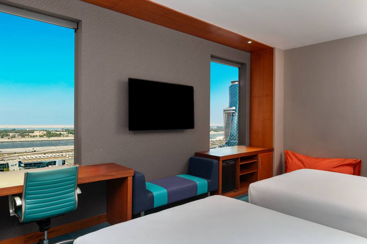 A hotel room with a large window, a desk, a television, a bed, and a seating area.