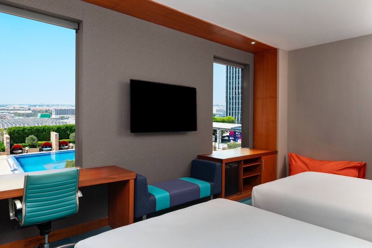 A hotel room with a bed, a desk with a chair, a TV mounted on the wall, and a view of a swimming pool and buildings.