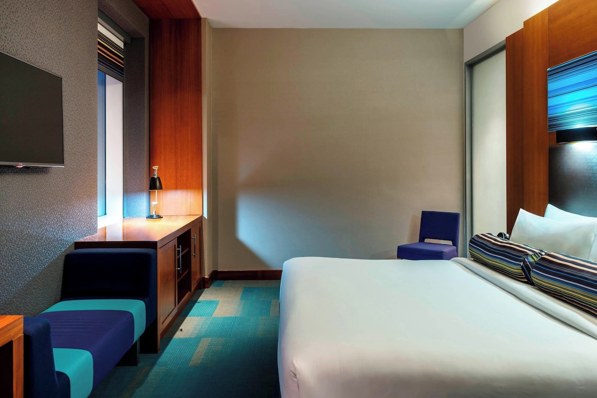 A hotel room with a bed, a desk with a lamp, a chair, and a television on the wall.