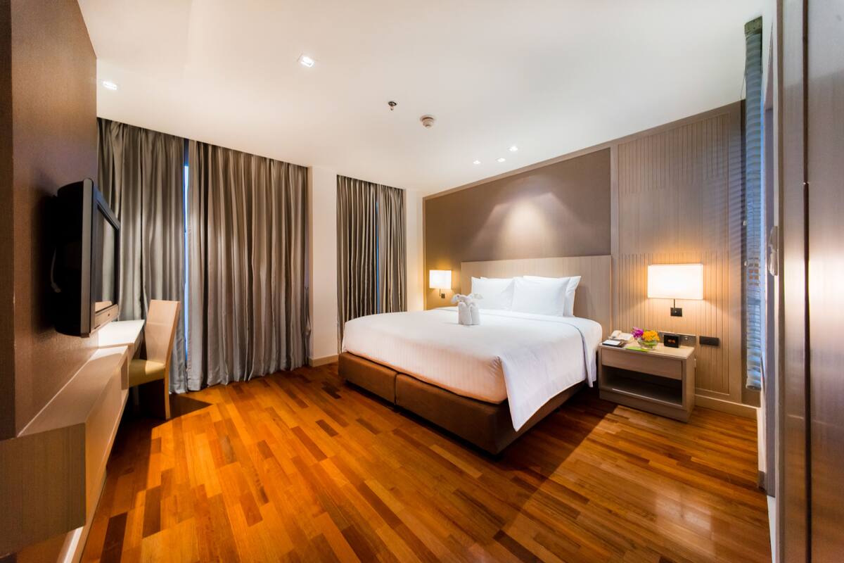 A hotel room with a large bed, wooden flooring, a flat-screen TV, a desk, and a nightstand with a lamp.