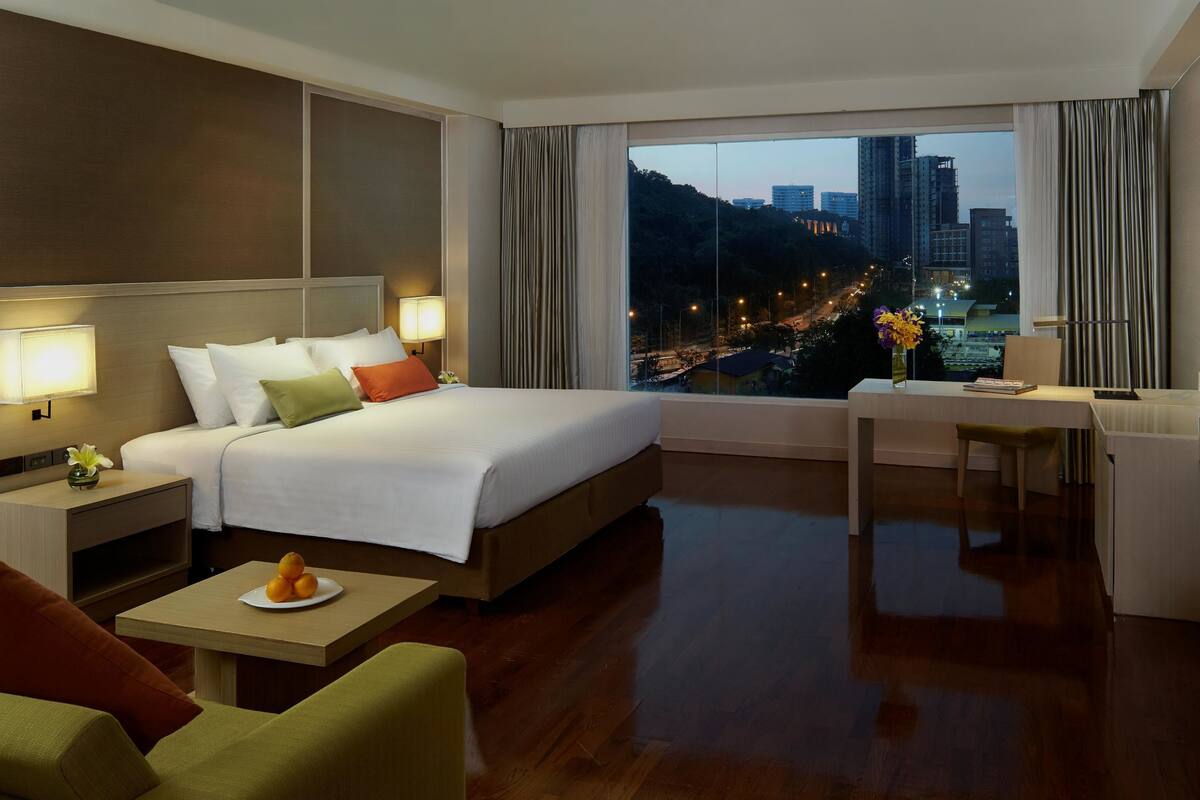 A hotel room with a bed, desk, TV, and a view of a cityscape at dusk.