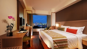 A hotel room with a large bed, a desk, a television, and a view of the city at dusk. - Signature Pattaya Hotel (Pattaya)