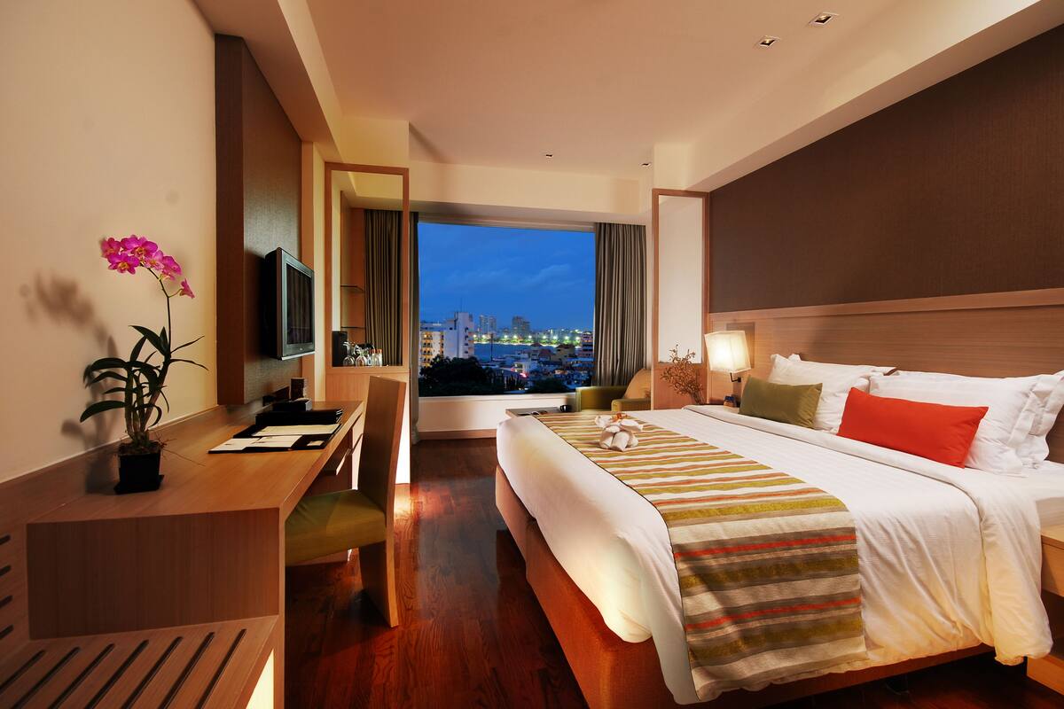A hotel room with a large bed, a desk, a television, and a view of the city at dusk.