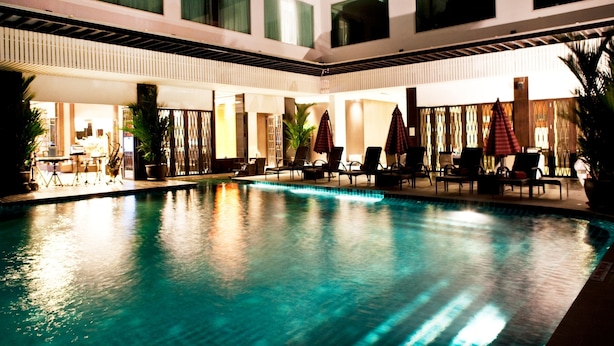 An indoor swimming pool with lounge chairs and umbrellas, surrounded by a modern building with large windows. - Signature Pattaya Hotel (Pattaya)