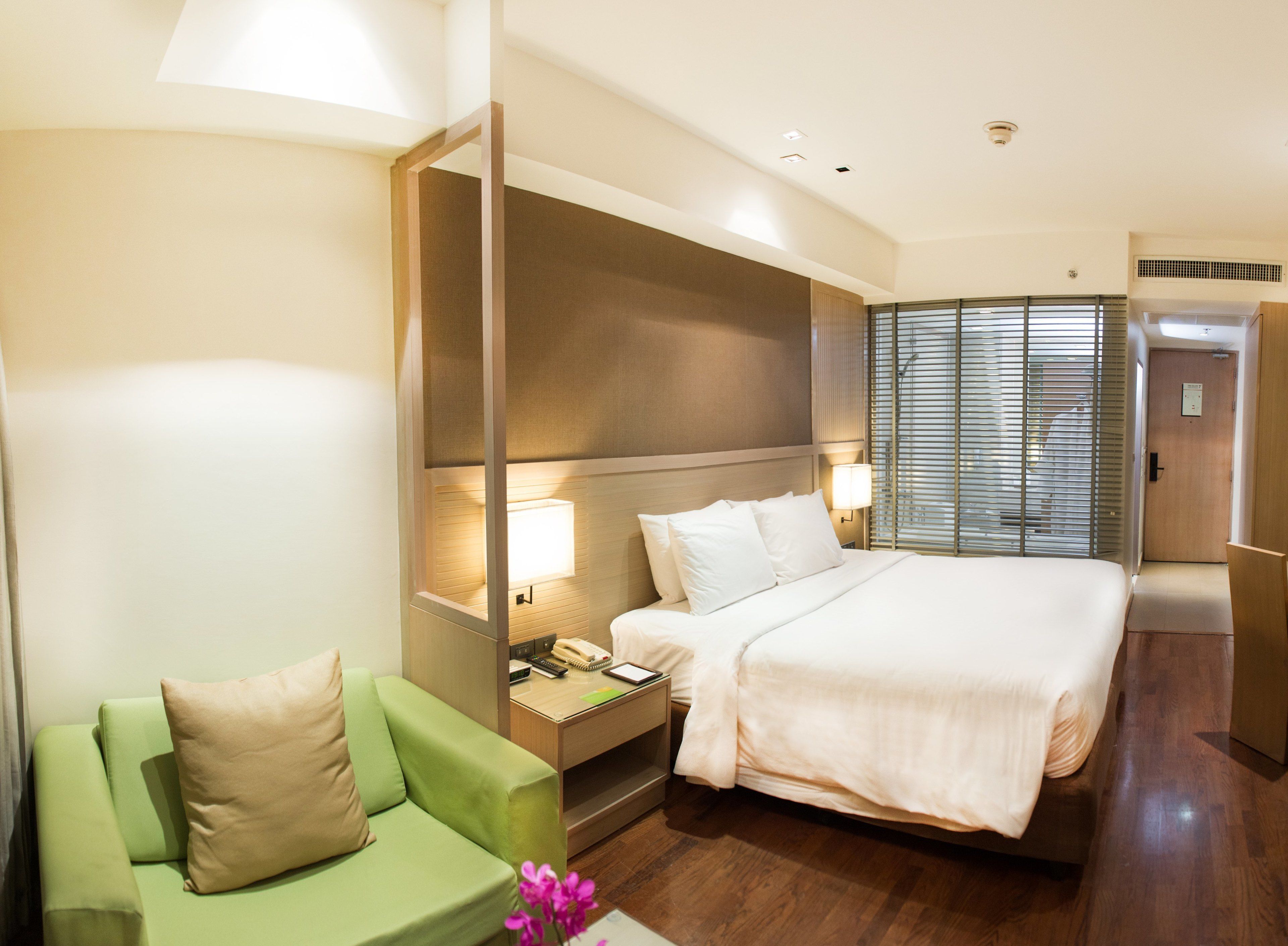 A hotel room with a bed, a green armchair, a nightstand with a lamp, and a window with blinds.