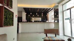 Zona de recepción de hotel con diseño moderno, que incluye un mostrador de recepción, lámparas colgantes y un área de descanso con cojines estampados.