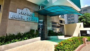 Entrada de un hotel con un letrero que dice "Hotel Bahia Suites", un acceso cubierto y un camino empedrado que lleva a la entrada.