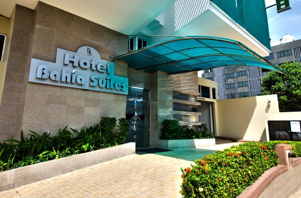Entrada de un hotel con un letrero que dice "Hotel Bahia Suites", un acceso cubierto y un camino empedrado que lleva a la entrada.