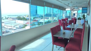 Un restaurante moderno con amplios ventanales que ofrecen vistas a la ciudad, asientos tapizados en rojo y un piso de baldosas impecable.
