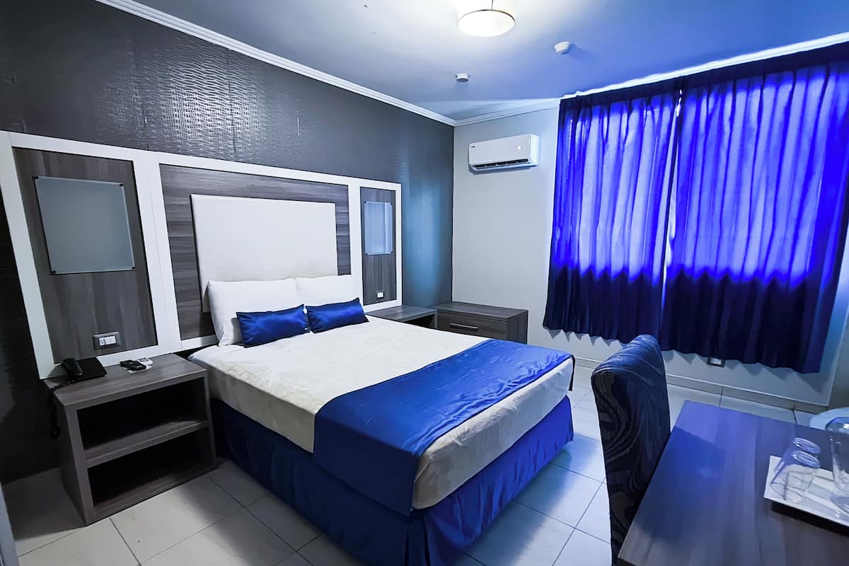 A hotel room with a large bed, a desk, a chair, a nightstand, a mirror, and an air conditioning unit.