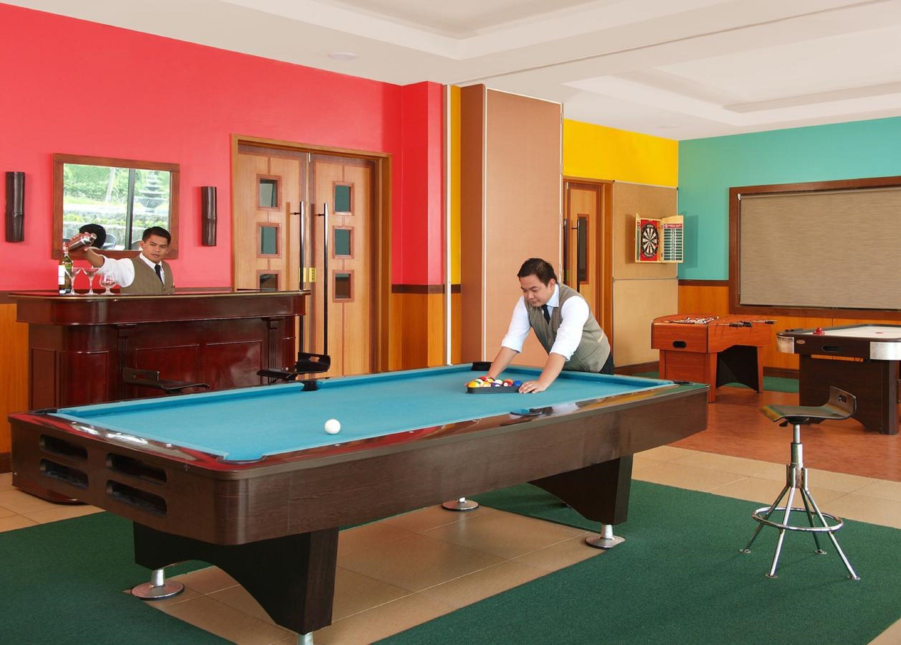 A pool table in a room with a bartender, a dartboard, and a billiards table.