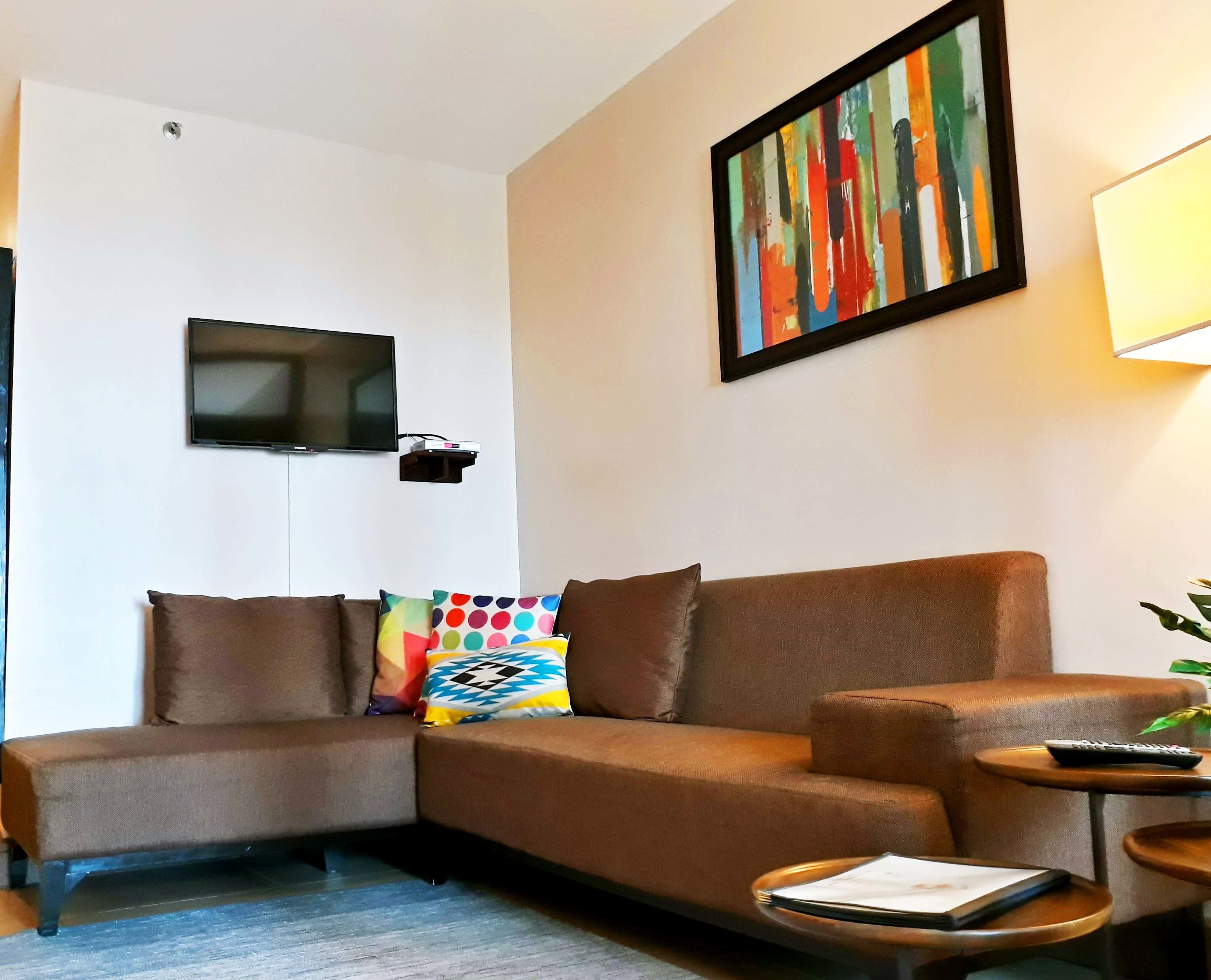 A modern living room with a brown sofa, a flat-screen TV mounted on the wall, a colorful abstract painting, and a small round table with a remote control.