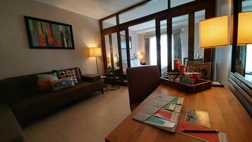 Habitación de hotel con un escritorio de madera, un sofá marrón con cojines de colores, un cuadro en la pared y un ventanal con cortinas.