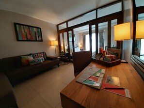 A hotel room with a wooden desk, a brown sofa with colorful pillows, a painting on the wall, and a large window with curtains. - Hotel Kimberly Tagaytay (Tagaytay)