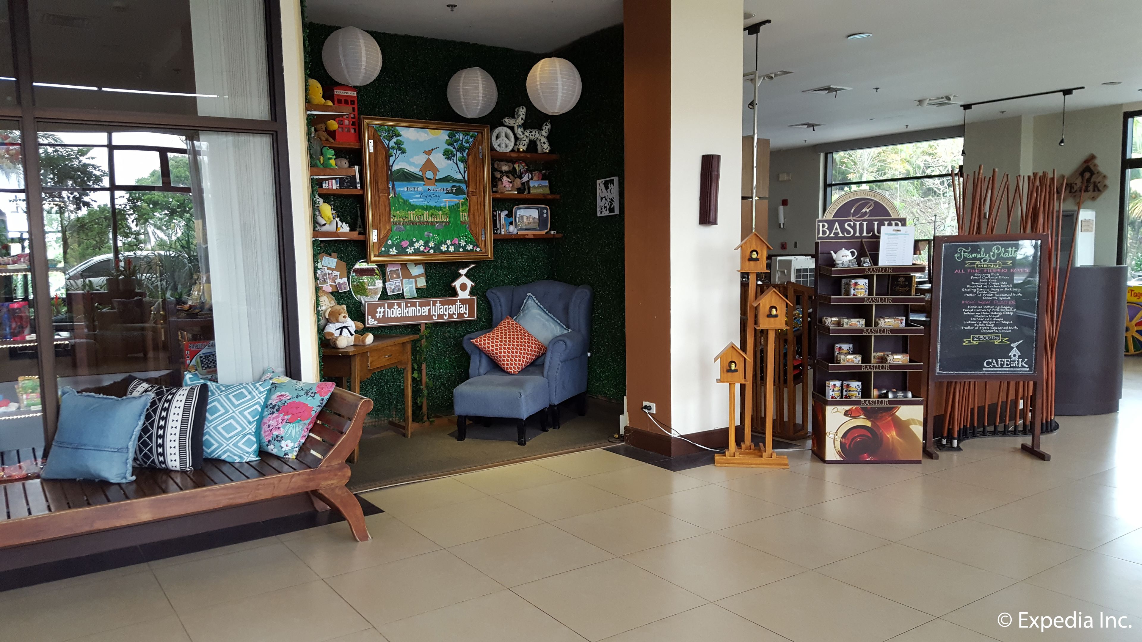 Un intérieur chaleureux avec un banc en bois, un fauteuil bleu et un mur décoré de divers objets, notamment un tableau encadré, un ours en peluche et une pancarte avec le hashtag #hotelkimberlytagay.