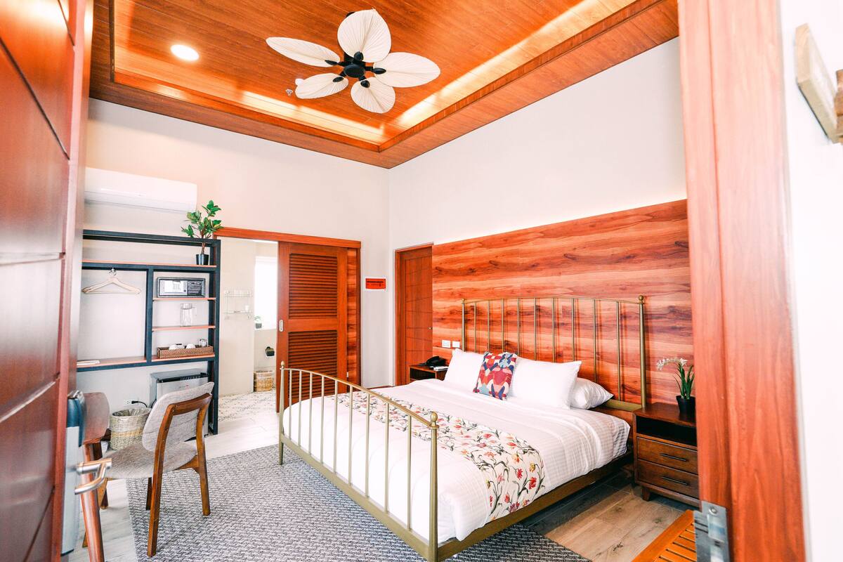 Une chambre moderne avec une tête de lit en bois, un grand lit avec une literie blanche, une table de chevet avec une lampe et un ventilateur de plafond.