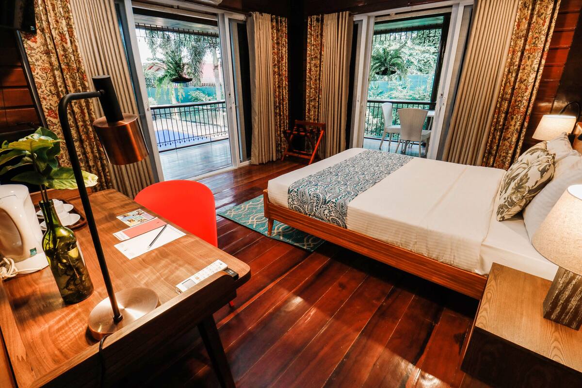 Une chambre d’hôtel avec un plancher en bois, un lit avec une literie à motifs, un bureau avec une lampe, une chaise et une vue sur un balcon équipé d’une chaise et d’une table.
