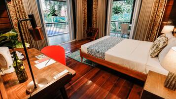 A hotel room with a wooden floor, a bed with patterned bedding, a desk with a lamp, a chair, and a view of a balcony with a chair and a table.