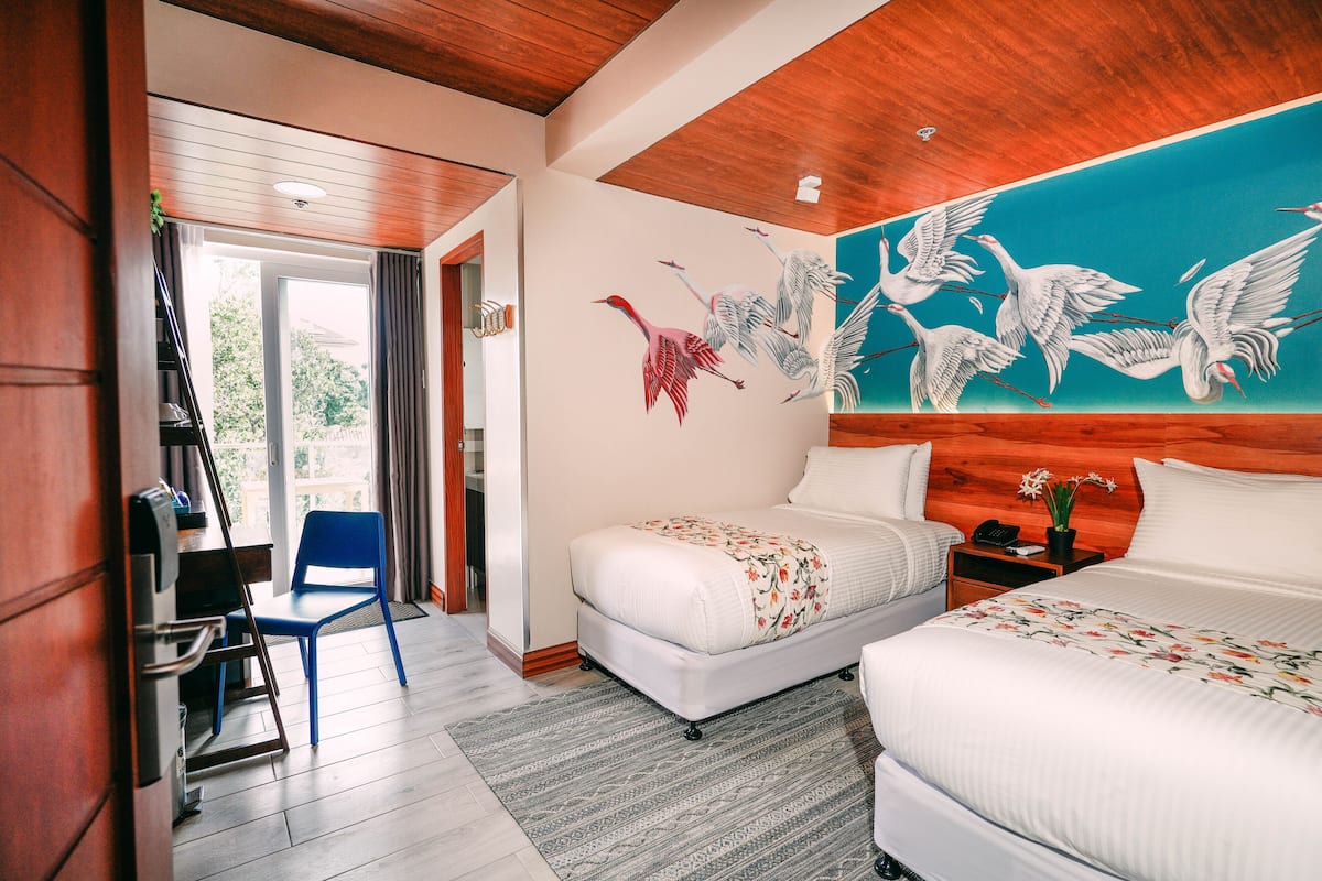 Une chambre d’hôtel avec deux lits, un plafond en bois, une fresque représentant des oiseaux, un bureau et une chaise.