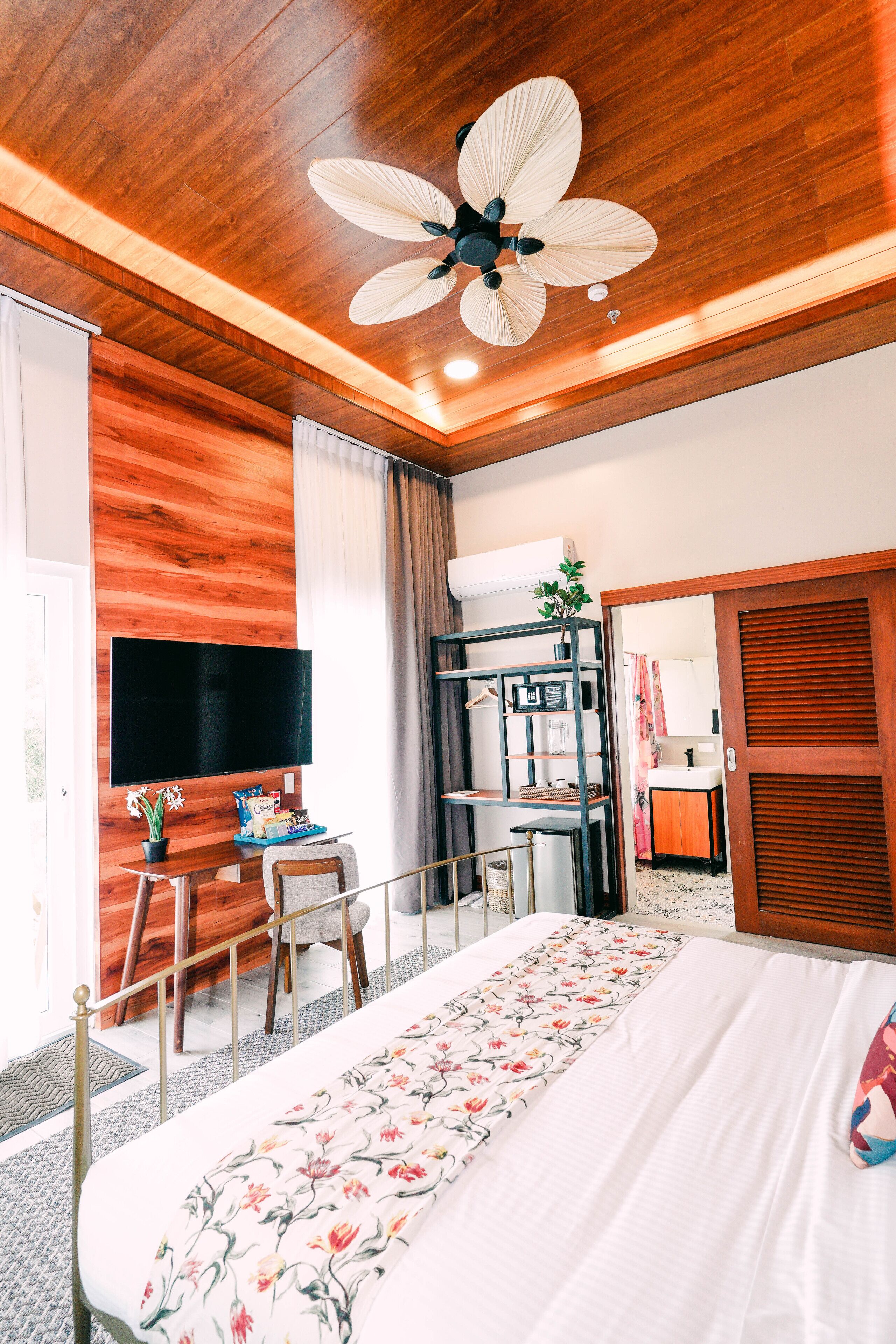 A hotel room with a wooden ceiling, a bed with a floral bedspread, a desk with a chair, a television, and a wardrobe.