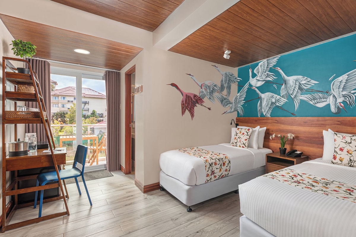 A hotel room with two beds, a wooden desk, a chair, a balcony with a view, and a mural of birds on the wall.