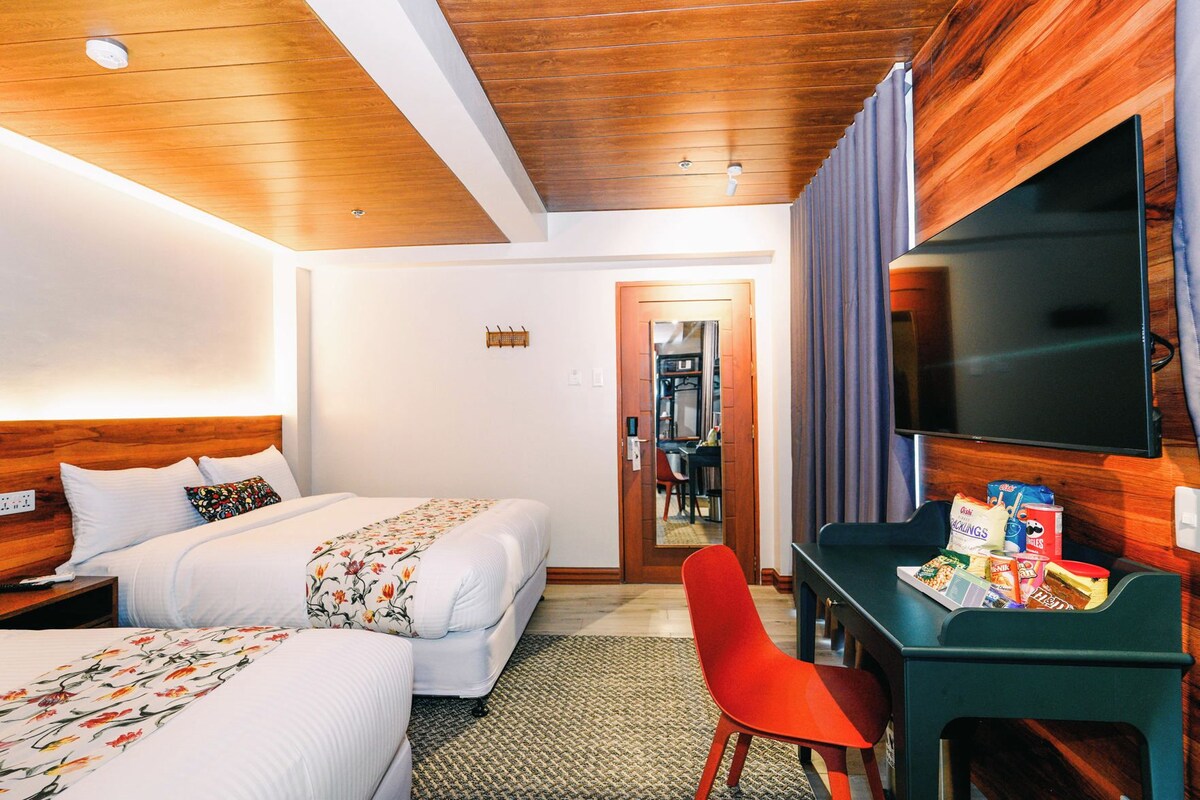 A hotel room with two beds, a desk with a chair, a television, and a small kitchenette area.