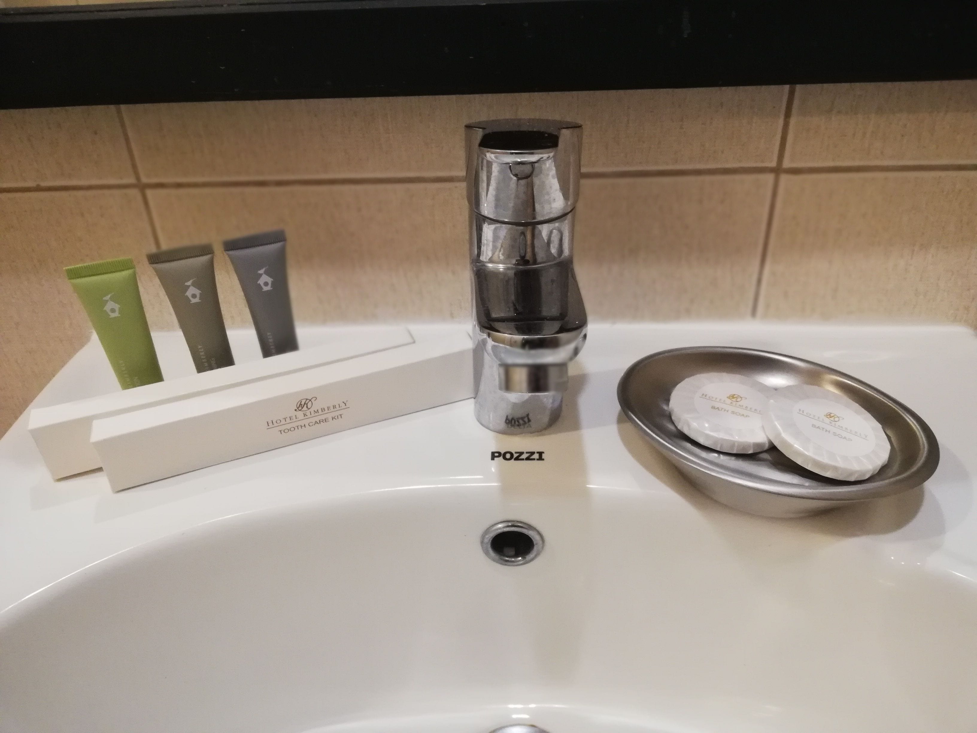 A hotel bathroom sink with a chrome faucet, a soap dish with three bars of soap, and a box of Hotel Kimberly Tooth Care Kit.