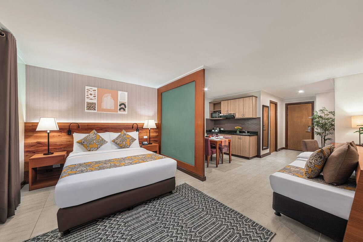 A hotel room with a bed, bedside tables, a desk, a chair, a small kitchen area, and a bathroom.