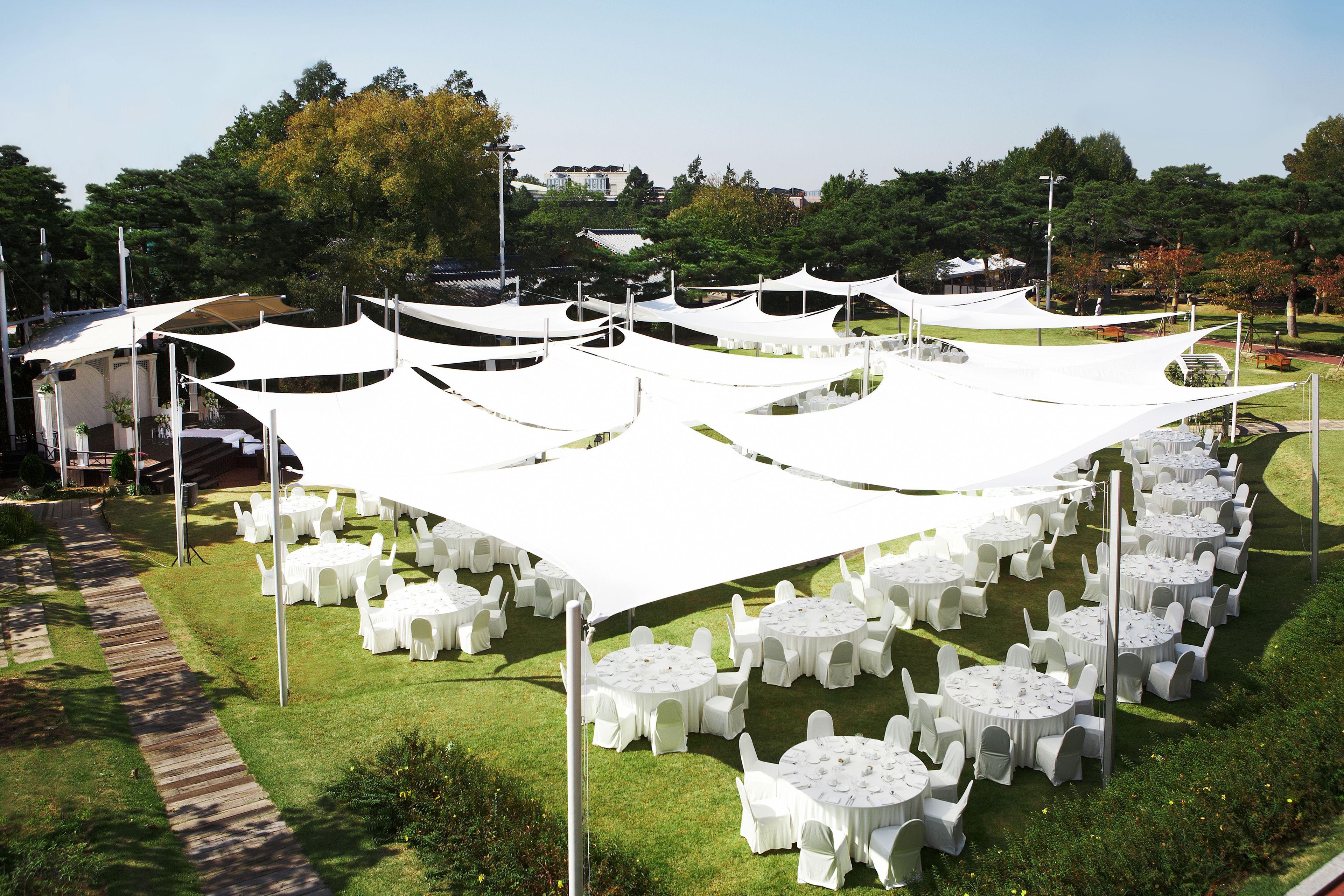 A garden setting with round tables and chairs under white canopies, surrounded by greenery and trees.